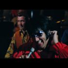 Ein Mann in einem roten Hemd, lächelt, hält ein Mikrofon, trägt Sonnenbrille. Zwei Musiker spielen im Hintergrund.