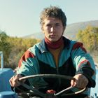 Keine sichtbaren Texte. Ein junger Mann fährt einen blauen Traktor in einer ländlichen Gegend, Bäume im Hintergrund.