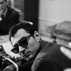 Ein Mann mit Sonnenbrille blickt über die Schulter, während im Hintergrund unscharf eine Person an einem Tisch sitzt.