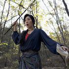 Eine Frau im Wald hält einen Stock hoch und ein aufgeschlagenes Buch in der Hand, ihr Ausdruck wirkt überrascht.