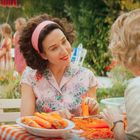 Zwei Frauen sitzen an einem Tisch im Freien, schälen Karotten. Im Hintergrund spielen Kinder. Es ist eine idyllische Gartenszene.