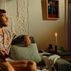 Ein Mann in gemusterter Kleidung sitzt auf einem Sofa. Ein anderer in einem Handtuch lehnt in einer Tür. Eine Kerze brennt.