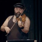 Ein bärtiger Mann spielt konzentriert Geige und trägt ein schwarzes Tanktop sowie eine Schirmmütze.