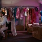 Eine Person schminkt sich an einem beleuchteten Spiegel. Bunte Kostüme hängen an einer Stange.