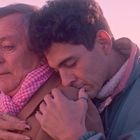 Zwei Personen umarmen sich in einer rosa getönten Landschaft bei Sonnenuntergang, eine trägt einen rosa Schal.