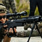Eine Person in Tarnkleidung zielt durch ein Zielfernrohr eines Gewehrs. Sie trägt Kopfhörer und eine Sonnenbrille.