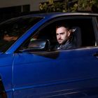 Der Text auf einem Autositz lautet "sparco". Ein Mann sitzt im Auto, schaut ernst und trägt eine schwarze Lederjacke.