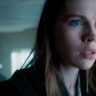 Eine Frau mit langen Haaren und blauen Augen schaut überrascht in die Ferne, im Hintergrund verschwommene Gestalt.