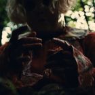 Eine schemenhafte Gestalt in einem Wald, die Hände nach vorne ausgestreckt, bekleidet mit einem zerzausten roten Anzug.