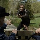 Keine Texte identifiziert. Eine angespannte Szene zeigt einen Mann mit gezogener Waffe und drei uniformierte Personen auf einem Lastwagen.