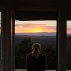 Sonnenuntergang durch ein großes Fenster, Person im Vordergrund, blickt auf bewaldete Hügel und untergehende Sonne.