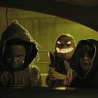Drei Kinder in gruseligen Kostümen stehen bei Nacht vor einem Autofenster; eines trägt eine Maske mit leuchtendem Lächeln.