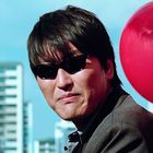 Rote Ballons mit teils erkennbaren weißen Logos. Ein Mann in Sonnenbrille trägt Anzug, vor einem unscharfen Gebäudehintergrund.