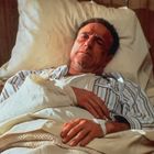 Ein Mann liegt in einem Bett, trägt gestreifte Pyjamas. Sein linker Arm ist bandagiert und ruht auf einer Decke.