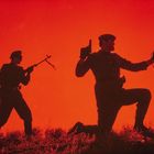 Zwei Soldaten-Silhouetten. Einer kniet, hält ein Fernglas und eine Pistole. Der andere steht mit einem Gewehr. Hintergrund rot.