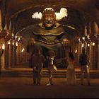 Eine massive Statue mit roten Augen und dampfenden Ohren sitzt in einer beleuchteten Höhle, umgeben von vier Personen.