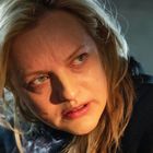 Es sind keine Texte sichtbar. Eine Frau mit blondem Haar schaut aufmerksam zur Seite, in einem gedämpften Licht.