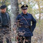 Ein Mann in einer dunklen Lederjacke mit Kreuzkette und ein Polizist in Uniform stehen im Wald.