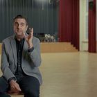 Keine sichtbaren Texte. Eine Person in einem Raum mit Bühne, die dunkelroten Vorhänge hat.