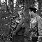 Ein junger Mann mit Medaillen spricht im Wald mit einer Frau in Militäruniform, die ein Gewehr und eine Decke trägt.