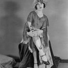 Dorothy Devore. Eine Frau in einem Kleid mit Hut sitzt, zeigt ihr Bein mit einer großen Schleifentätowierung.