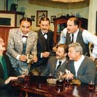 Rahmen mit Text: „Hallen Stimmen in alter Wollt, möge er dich stimmen der treue Kolb.“ Sechs Männer in Anzügen in einem Raum.