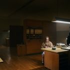 Es gibt keinen sichtbaren Text. Eine Person sitzt in einem Büro bei einem Schreibtisch, eine andere steht daneben.