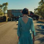 Eine Person in einem blauen Kleid steht auf einer leeren Straße, vor einem Lkw-Unfall, umgeben von Bäumen und Trümmern.