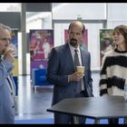 „Schlager“ auf plakat im hintergrund. Drei personen stehen an einem tisch, trinken kaffee, im modernen gebäuderaum.