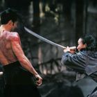 Zwei Personen mit Schwertern im Duell, eine trägt einen Regenmantel, die andere ist oberkörperfrei mit tätowiertem Rücken.