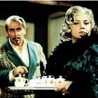 Ein Mann mit Schnurrbart und ein Frau mit Locken tragen elegante Kleidung. Sie hält ein Tablett mit Teetassen.