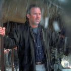 Ein Mann steht unter einem Zelt im Regen, hält einen Stock und trägt eine Jacke. Im Hintergrund unscharf arbeitende Personen.