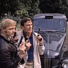 Zwei Männer mit Pistolen stehen vor einem alten, dunklen Auto mit dem Schriftzug "Austin" auf der Motorhaube. Im Hintergrund Bäume.