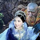 Eine lächelnde, elegant gekleidete junge Frau in Blau, mit aufwändigem Kopfschmuck. Neben ihr ein älterer, bärtiger Mann mit ungewöhnlicher Hautfarbe und Schmuck. Hintergrund zeigt eine düstere Waldszene.