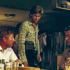 Dosen mit "OLYMPIA BEER"-Logo. Drei Männer in einem Raum, Kleidung und Getränkedosen sind sichtbar. Warmes Licht.