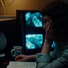 Regale beschriftet mit „ZONE KEYS“, Überwachungsmonitore, eine Frau liest, Ventilator und eine Tasse mit einem Logo.