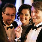 Ich kann den Text nicht erkennen. Drei Personen in eleganter Kleidung lächeln bei einer fröhlichen Feier.