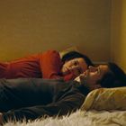 Eine Frau in Rot und ein Mann in Dunkelgrau liegen nebeneinander auf einem Bett und schauen ruhig.