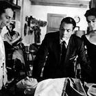 Vier Menschen, davon drei stehend um eine aufgebahrte Person. Hintergrund möbliert, Mann raucht Pfeife. Schwarz-weiß.