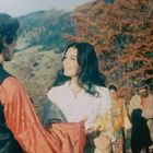 Zwei Menschen in traditioneller Kleidung tanzen draußen vor Herbstbäumen, umgeben von anderen Personen.