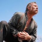 Ein Mann kniet auf dem Boden, presst ein blutiges Tuch gegen seinen Bauch. Der Himmel ist klar und blau im Hintergrund.