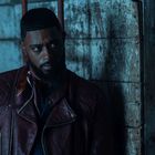 Ein Mann mit Bart und Lederjacke lehnt an einer verwitterten Holzwand, blickt ernst zur Seite.