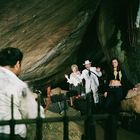 Drei Personen in einer Höhle, die Hände erhoben. Eine Frau steht im Vordergrund, ein Mann schaut mit einer Waffe zu.