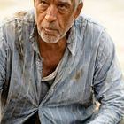 Ein älterer Mann mit grauem Haar trägt ein zerrissenes Hemd und hat eine mit Stoff umwickelte Hand. Sein Ausdruck ist ernst.