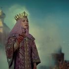 Eine Frau in edlem Gewand und Krone steht vor verschwommener, märchenhafter Architektur.