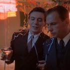 Zwei Männer in Anzügen stehen auf einer Feier mit bunten Luftschlangen und trinken, umgeben von warmem, gedämpftem Licht.