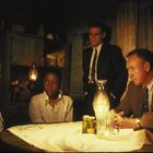 Vier Personen sitzen und stehen um einen Tisch mit einer Öllampe. Die Stimmung wirkt angespannt und ernst.