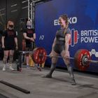 „Scottish Powerlifting“ links, „British Powerlifting“ rechts. Frau hebt Hantel, Zuschauer und Betreuer um sie herum.