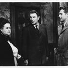 Drei Personen in formeller Kleidung stehen in einem Raum mit verziertem Hintergrund. Sie blicken ernst.