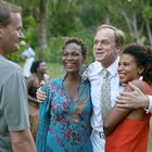 Vier Menschen stehen lachend zusammen im Freien, ein Mann umarmt zwei Frauen. Im Hintergrund sind Bäume.